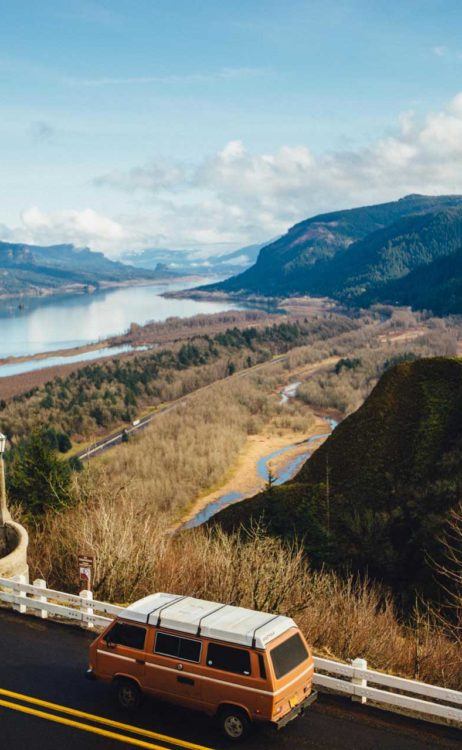 orange vintage camper van driving a paved highway looking over an open landscape with tree-covered hills and a large river