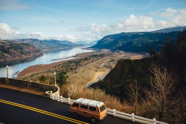 orange vintage camper van driving a paved highway looking over an open landscape with tree-covered hills and a large river